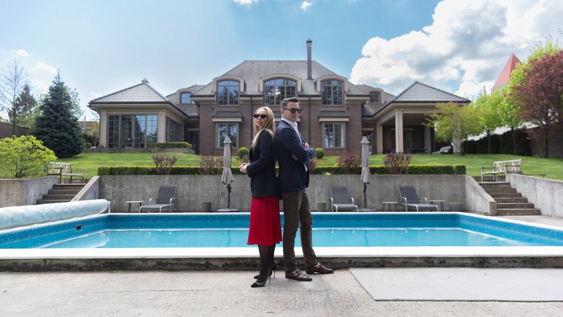 A man and a woman standing back to back. They are in front of a large home and pool.