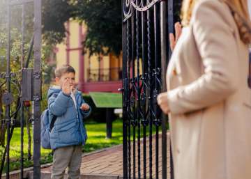 A child standing at a gate and waving goodbye to his mother.