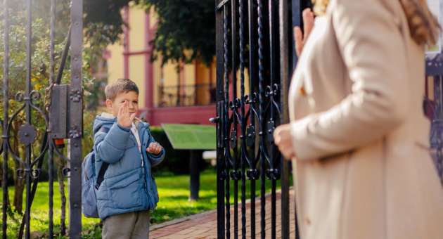 A child standing at a gate and waving goodbye to his mother.