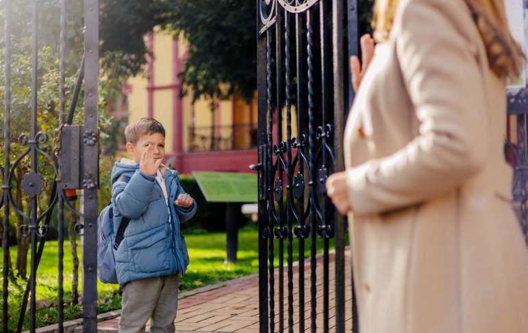 A child standing at a gate and waving goodbye to his mother.