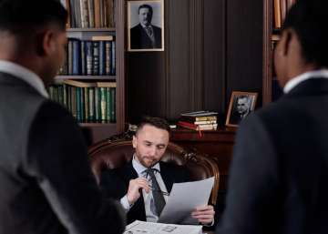 A lawyer sits in his office chair. Two other lawyers stand at the other side of his desk.