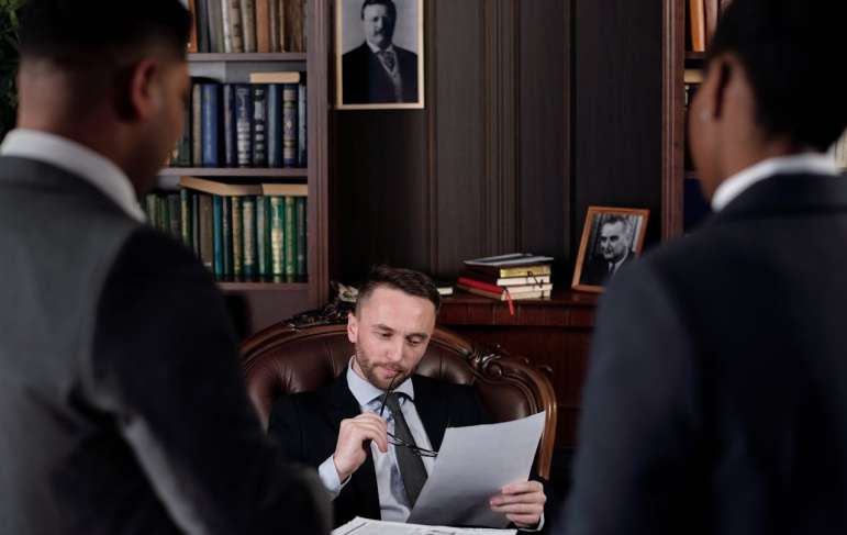 A lawyer sits in his office chair. Two other lawyers stand at the other side of his desk.