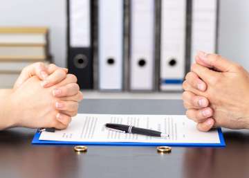 Two people sitting across from each other. They have their hands folded on a table. There are documents and rings placed between the two.