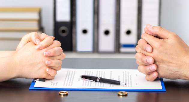Two people sitting across from each other. They have their hands folded on a table. There are documents and rings placed between the two.