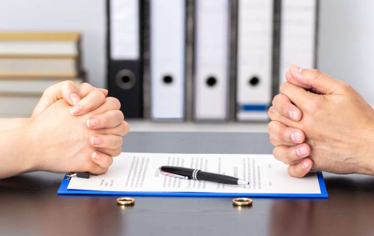 Two people sitting across from each other. They have their hands folded on a table. There are documents and rings placed between the two.